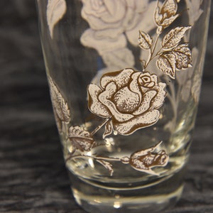 K&ouml;nnte beinhalten: Ein klares Glas mit einem wei&szlig;en und braunen Blumendesign. Das Design zeigt Rosen und Bl&auml;tter.