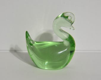 May include: A decorative glass swan figurine. The swan is primarily a translucent green color with a clear head and neck. The swan has a curved neck and a detailed beak and tail.