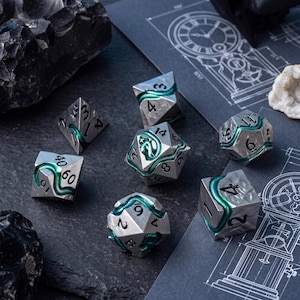 May include: A set of seven polyhedral dice made of silver-toned metal with a textured surface. Each die features a winding, vibrant green snake relief that snakes across the faces, separating the black engraved numbers on each geometric shape.