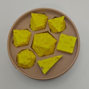 Könnte beinhalten: Ein Set aus sechs gelben polyedrischen Würfeln mit einer strukturierten Oberfläche, die an Schweizer Käse erinnert, angeordnet in einem runden beigen Tablett. Die Würfel umfassen verschiedene geometrische Formen wie W20, W12, W10, W8, W6 und W4 mit geprägten Zahlen.