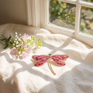 Puede incluir: Broche de libélula con cuerpo dorado y alas adornadas con piedras preciosas rosas y cristales transparentes. Los ojos de la libélula son pequeñas piedras verdes. La broche está sobre una tela blanca.