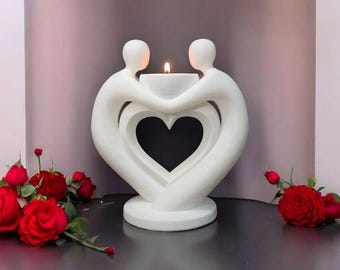 May include: A white candle holder in the shape of two figures embracing, forming a heart. A lit candle sits in the center. Red roses are arranged on either side of the candle holder.