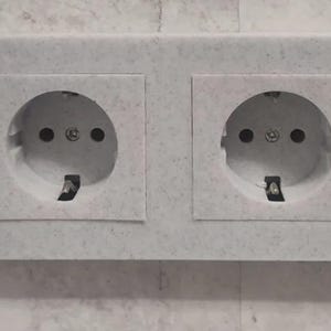 May include: A double electrical outlet with two round sockets. The sockets are gray with two small holes and a grounding pin. The outlet is set in a light gray, textured faceplate.