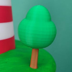 Może przedstawiać: Miniaturowa scena wydrukowana w 3D przedstawiająca zielone drzewo z brązowym pniem na zielonej podstawie, obok czerwono-białej latarni morskiej. Tło jest w jednolitym turkusowym kolorze.