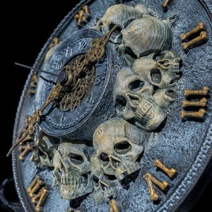 Könnte beinhalten: Eine gotische Uhr mit einem dunkelgrauen Zifferblatt und goldenen römischen Ziffern. Die Uhr hat ein detailliertes Design von Totenköpfen um das Zifferblatt und verzierte goldene Zeiger. Das Zifferblatt der Uhr hat einen Durchmesser von etwa 25 cm.