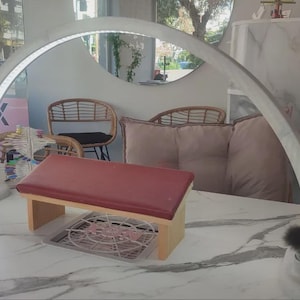 Puede incluir: Una configuración de salón de uñas con un reposabrazos acolchado rojo, una lámpara LED curva y una superficie con diseño de mármol. La escena incluye un ventilador decorativo, un espejo redondo y productos para el cuidado de las uñas, creando un ambiente profesional y acogedor.