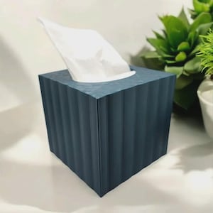 Puede incluir: Un dispensador de pañuelos cuadrado, azul oscuro, con detalles verticales acanalados. Los pañuelos blancos sobresalen de la abertura superior. La caja está sobre una superficie blanca, con plantas verdes en el fondo.
