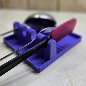 May include: A purple utensil rest holds a pink spatula, a metal spoon, and a black-handled utensil. The rest has a rectangular base with slots and vertical supports to hold the utensils. A stainless steel ladle is in the background.