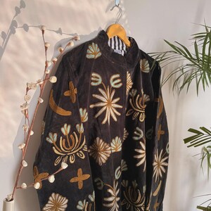 May include: A dark brown jacket with intricate floral embroidery in gold, light blue, and cream colors. The jacket is hanging on a wooden hanger against a white wall. The embroidery features various flower and leaf designs.