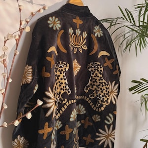 May include: A black velvet jacket with elaborate embroidery. The jacket features gold, white, and turquoise floral and geometric designs, along with two leopard figures. The jacket is hanging on a wooden hanger.