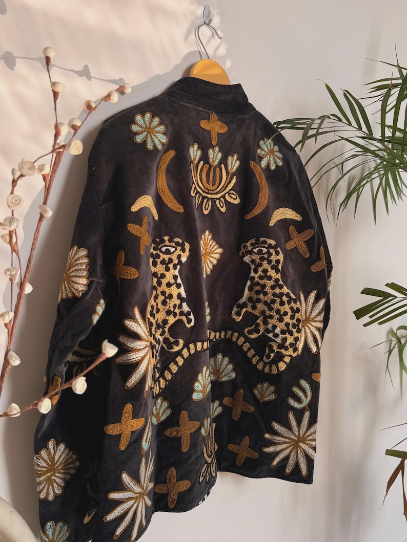 May include: A black velvet jacket with elaborate embroidery. The jacket features gold, white, and turquoise floral and geometric designs, along with two leopard figures. The jacket is hanging on a wooden hanger.