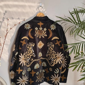 May include: Black jacket with gold and cream embroidery. The jacket features two leopard designs and floral and cross patterns. The jacket is hanging on a wooden hanger.
