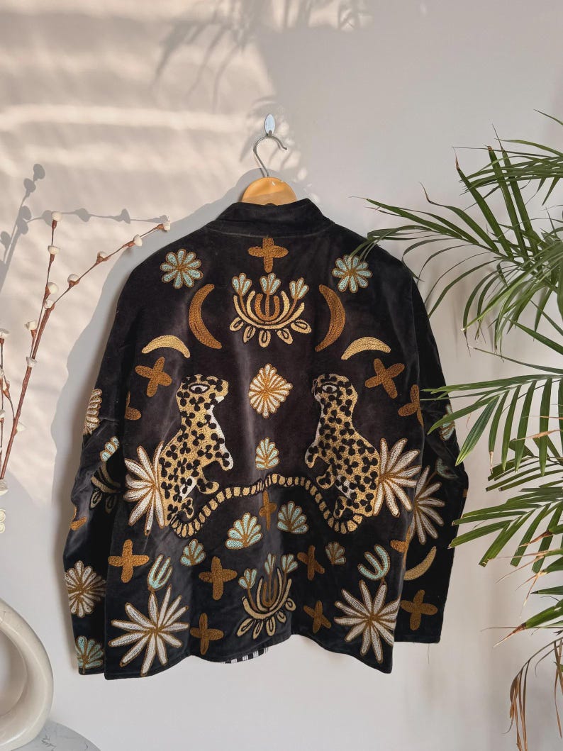 May include: Black jacket with gold and cream embroidery. The jacket features two leopard designs and floral and cross patterns. The jacket is hanging on a wooden hanger.