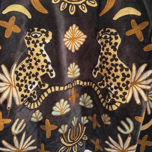 May include: Close-up of a dark gray textile with intricate embroidery. The design features two leopard-like figures with spotted patterns, surrounded by floral and geometric motifs in gold, cream, and teal. The overall aesthetic is bohemian and artistic.
