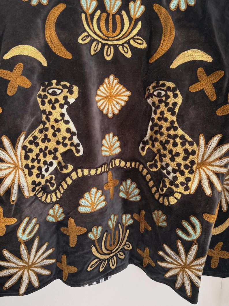 May include: Close-up of a dark gray textile with intricate embroidery. The design features two leopard-like figures with spotted patterns, surrounded by floral and geometric motifs in gold, cream, and teal. The overall aesthetic is bohemian and artistic.