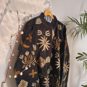 May include: A dark brown jacket with intricate floral embroidery in gold, light blue, and cream colors. The jacket is hanging on a wooden hanger against a white wall. The embroidery features various flower and leaf designs.