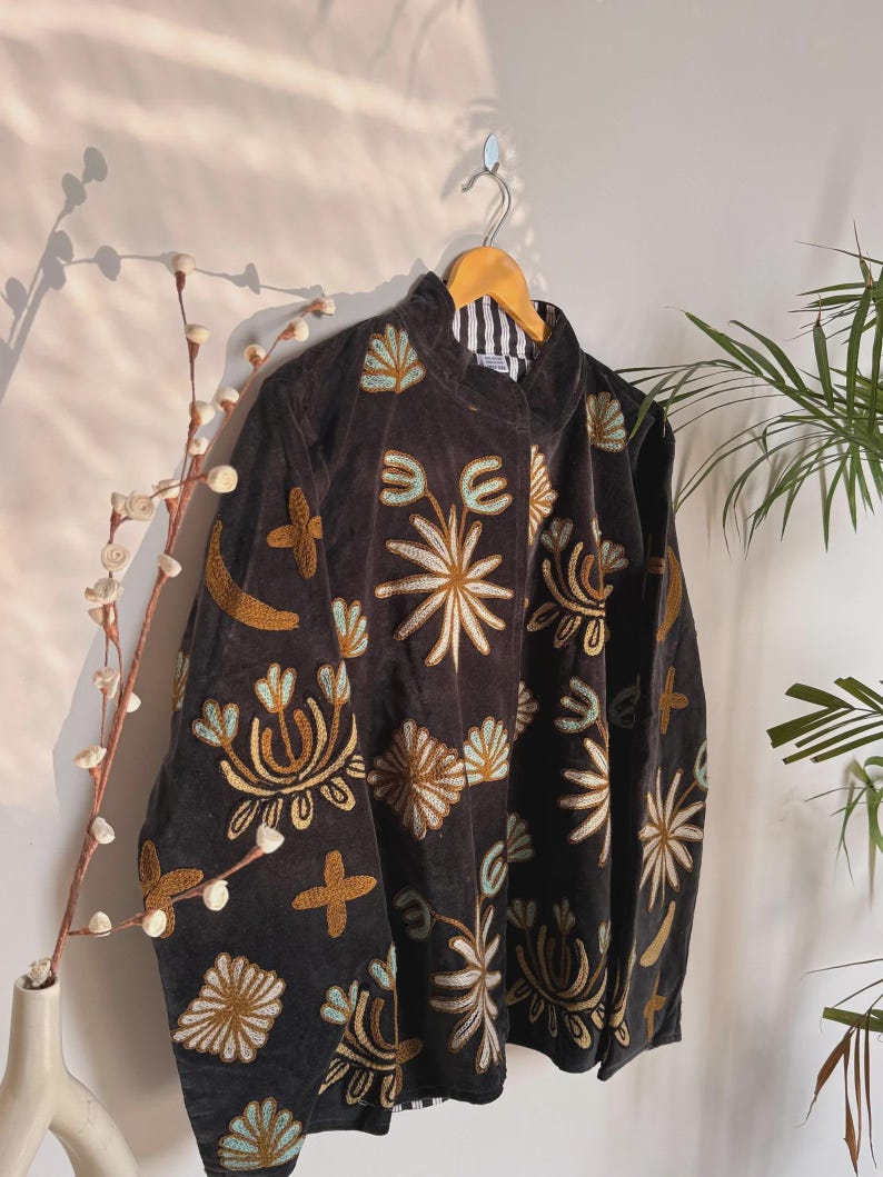 May include: A dark brown jacket with intricate floral embroidery in gold, light blue, and cream colors. The jacket is hanging on a wooden hanger against a white wall. The embroidery features various flower and leaf designs.