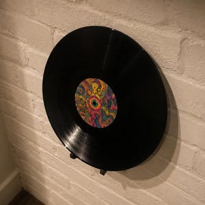 May include: A black vinyl record with a colorful, psychedelic label. The record is mounted on a black stand against a white brick wall. The label features a vibrant design with swirling patterns and a central eye-like motif.