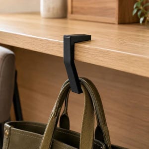 Könnte beinhalten: Ein mattschwarzer Schreibtischhaken aus Metall mit L-förmigem Design, der an die Kante einer Holzoberfläche geklemmt wird. Der Haken verfügt über einen nach unten gebogenen Arm, der die Griffe einer olivgrünen Segeltuchtasche sicher hält.