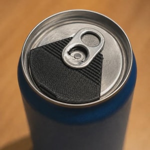 Könnte beinhalten: Eine blaue Aluminium-Getränkedose mit einem silbernen Metall-Zugringdeckel. Eine schwarze, texturierte, geometrische Kunststoffabdeckung ist auf der Oberseite der Dose angebracht und verdeckt teilweise die Oberfläche des Deckels.