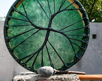 May include: A circular frame drum with a translucent green drumhead featuring a black cord lacing pattern that resembles a tree. The drum is accompanied by a mallet with a grey fabric-covered head and a wooden handle, positioned in front of the drum.