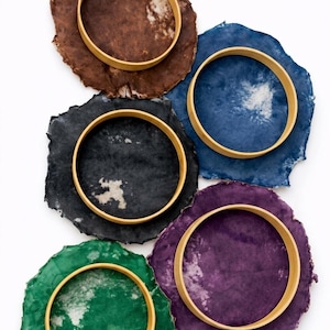 May include: An overhead shot displays six colorful, irregularly shaped coasters. Each coaster is a different color: brown, blue, black, green, and purple. A gold-colored ring sits atop each coaster, creating a striking contrast.