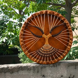 Pode incluir: Um tambor de moldura redondo com uma pele castanho-clara apresentando um design central de árvore tecido com cordões castanhos claros e escuros. O tambor tem uma borda de madeira espessa e texturizada, com cordões claros que irradiam do centro.