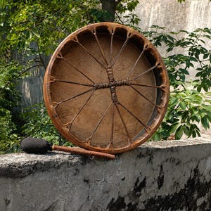 May include: A round wooden frame drum with a tan skin head stretched across the front. The drum features a network of thin leather lacing that connects the rim to a central cross-shaped handle. A wooden mallet with a black fabric head is included.