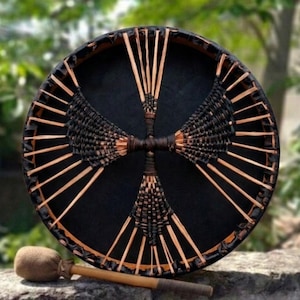 May include: A round, black drum with a woven design and wooden accents. The drum has a dark brown rim and is accompanied by a mallet with a tan head and a wooden handle. The drum is resting on a stone surface.