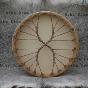 May include: A round, light brown drum with a natural hide surface and a tree-like design made of woven cord. The drum has the text "Atha Drum" repeated around the edge. The drum is set against a gray, textured background.