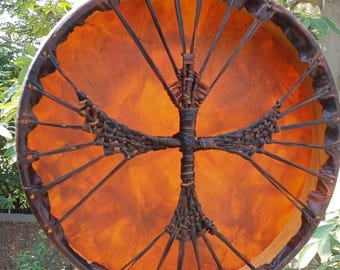 May include: A round, handcrafted drum with a warm orange center and a dark brown leather frame. The drum features a central woven design and radiating spokes, creating a visually striking instrument. The drum is likely used for music or spiritual practices.