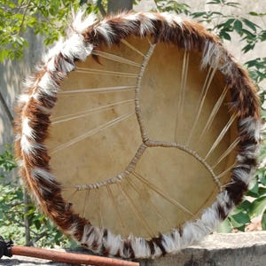 May include: A round, handmade drum with a natural hide surface and a decorative fur trim in shades of brown and white. The drum has a network of internal supports and a wooden handle.
