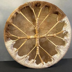 May include: A round, handcrafted drum with a natural hide head and a wooden frame. The drum's edge is trimmed with brown fur and white accents. The interior features a cross-shaped design made of woven material and wooden spokes.