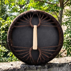 May include: A circular drum with a dark brown fur rim and a black central surface. It has a light brown wooden handle and is laced with brown cord. The background shows blurred green leaves and branches.