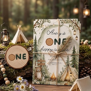 May include: Invitations with a camping theme, tied with twine. The text reads "Avery is ONE happy camper!" with illustrations of a bear, tent, and campfire. A wooden slice with "ONE" is also visible, alongside a small tent model.