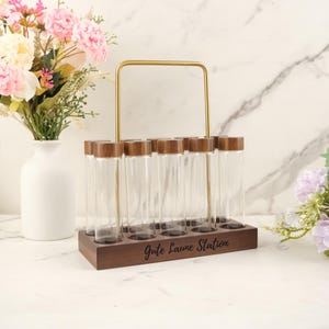 May include: A wooden spice rack with eight clear glass tubes, each capped with a wooden lid. The rack has a gold-colored handle and the words "Gute Laune Station" are printed on the front. A white vase with flowers is in the background.