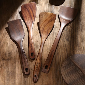 Könnte beinhalten: Eine Sammlung von Küchenutensilien aus Holz, darunter Pfannenwender und ein Löffel, mit graviertem Text. Die Utensilien sind in verschiedenen Brauntönen gehalten und auf einer Holzoberfläche angeordnet. Der Text enthält Sätze wie "Cook with Love".