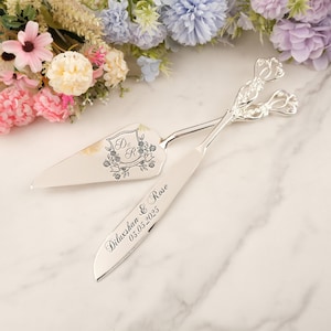 May include: Silver cake server and knife set. The server features a floral crest with the initials "D & R". The knife is engraved with "Diluxshan & Rose 05.05.2025". The handles have a decorative floral design. Perfect for weddings.