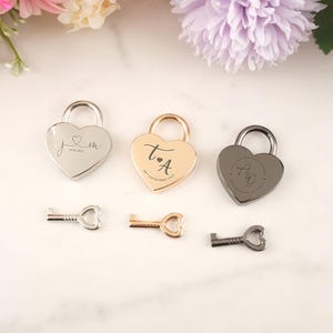 May include: Three heart-shaped padlocks in silver, gold, and gunmetal, each with a matching key. The padlocks are engraved with initials and dates. The background includes pink and purple flowers.