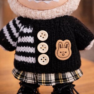Op de afbeelding: Een pluche pop met een zwarte trui met wit en zwart gestreepte mouwen, drie cr&egrave;mekleurige knopen en een kleine konijnenpatch. De pop heeft een geruite rok en zwarte schoenen. De kop van de pop is wit en pluizig.