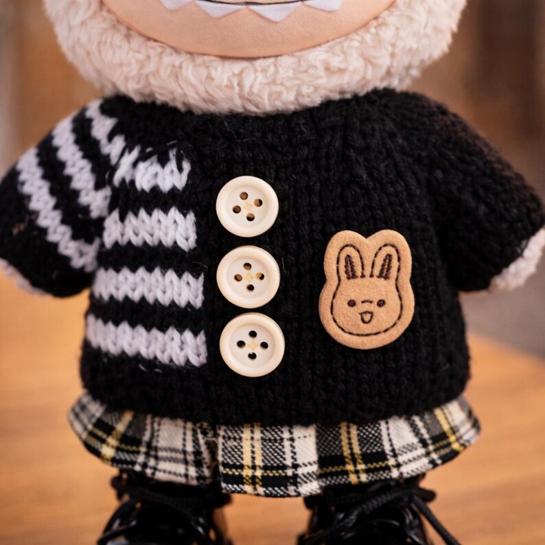 Op de afbeelding: Een pluche pop met een zwarte trui met wit en zwart gestreepte mouwen, drie cr&egrave;mekleurige knopen en een kleine konijnenpatch. De pop heeft een geruite rok en zwarte schoenen. De kop van de pop is wit en pluizig.