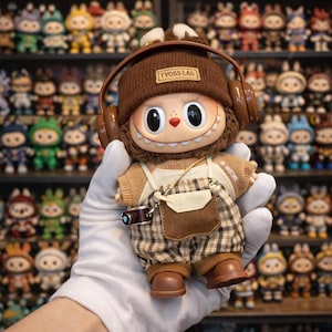 May include: A collectible doll with a brown knit cap and headphones, wearing a tan sweater and plaid overalls. The doll has large eyes and a small camera accessory. The text "TYOSS LAB" is on the cap.