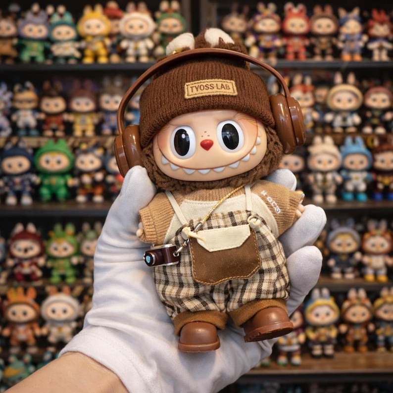 May include: A collectible doll with a brown knit cap and headphones, wearing a tan sweater and plaid overalls. The doll has large eyes and a small camera accessory. The text "TYOSS LAB" is on the cap.