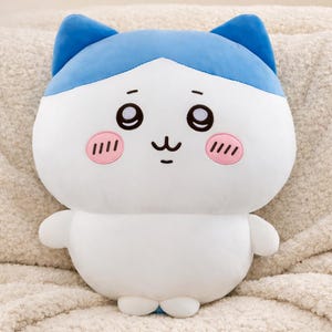 Puede incluir: Un peluche con forma de personaje de dibujos animados, con cuerpo blanco, orejas y cabeza azules y mejillas rosas. El personaje tiene ojos grandes y expresivos y una boca pequeña y sonriente. El peluche es suave y tierno.