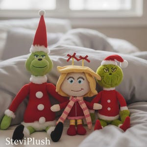 Può includere: Tre personaggi natalizi di peluche: un Grinch verde con costume e cappello da Babbo Natale rosso, una ragazza con i capelli biondi e una sciarpa rossa, e un altro Grinch con cappello da Babbo Natale. I Grinch hanno piedi neri e la ragazza scarpe rosse.