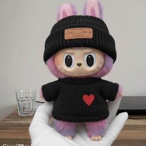 Pode incluir: Um brinquedo de pelúcia com um design único, com gorro e suéter pretos com um coração vermelho. O brinquedo tem um rosto claro, olhos grandes e pele roxa e rosa. Um pequeno patch de couro está no gorro.