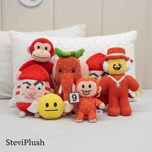 Pode incluir: Uma coleção de brinquedos de pelúcia em várias formas e cores. Os brinquedos incluem um macaco vermelho, um morango, uma carinha sorridente amarela com asas e uma figura com um chapéu. Os brinquedos estão dispostos em uma superfície branca.