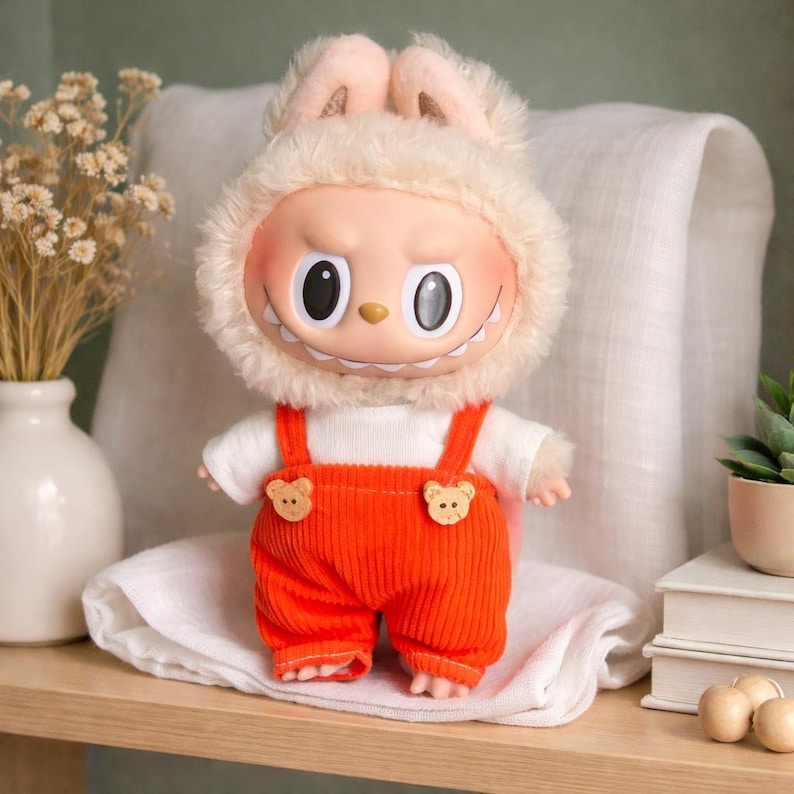 Peut inclure: Poup&eacute;e en peluche au design unique. Le visage clair de la poup&eacute;e pr&eacute;sente de grands yeux noirs et un sourire &eacute;dent&eacute;. Elle porte une capuche en fourrure avec des oreilles de lapin, une chemise blanche et une salopette en velours c&ocirc;tel&eacute; orange avec des boutons en forme d'ours. La poup&eacute;e est sur une surface en bois.