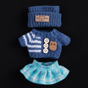 Op de afbeelding: Een blauwe gebreide poppenoutfitset, bestaande uit een muts, een trui en een geplooide rok. De trui heeft witte strepen, houten knopen en een berenvormige patch. De muts en trui hebben een label met "Doll Wishes".