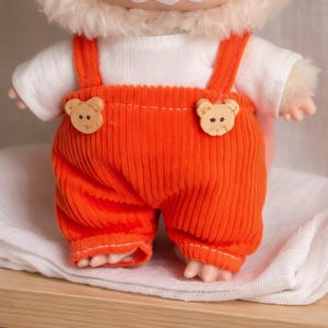 Peut inclure: Une poup&eacute;e v&ecirc;tue d'une salopette en velours c&ocirc;tel&eacute; orange avec des boutons en bois en forme d'ours et un t-shirt blanc. La salopette a des bretelles et arrive aux genoux. La poup&eacute;e a une t&ecirc;te poilue et de petites mains et pieds.